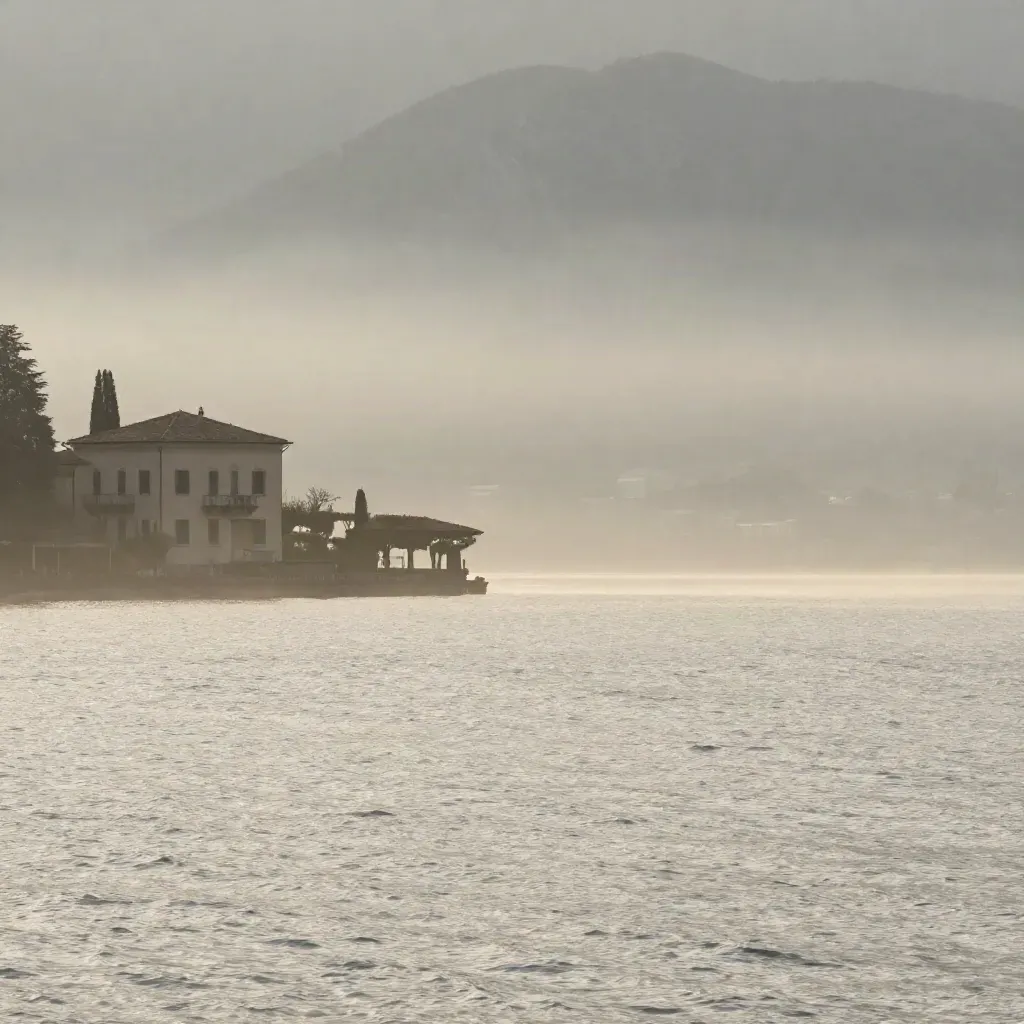 Lago di Como all'alba con montagne nella foschia e acqua argentea sotto.